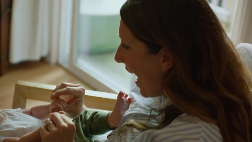 A smiling mother cradles her newborn baby in her lap and tenderly plays with his hands