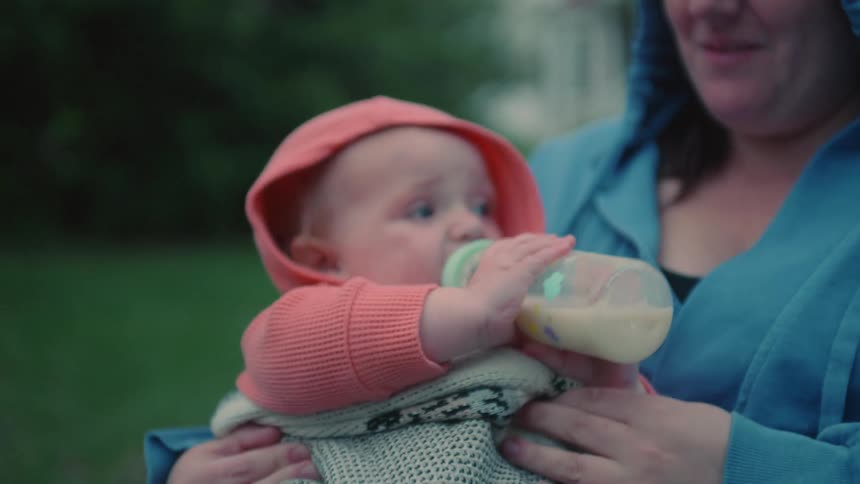 Close up of a happy baby in a hooded sweatshirt drinking milk from a bottle in its mother's arms