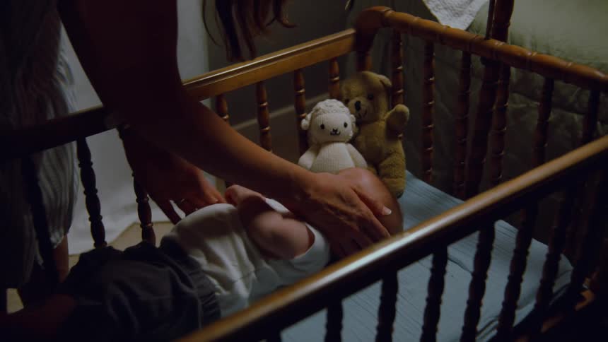 A mother gently soothes her newborn baby lying awake in a wooden crib in a dimly lit bedroom