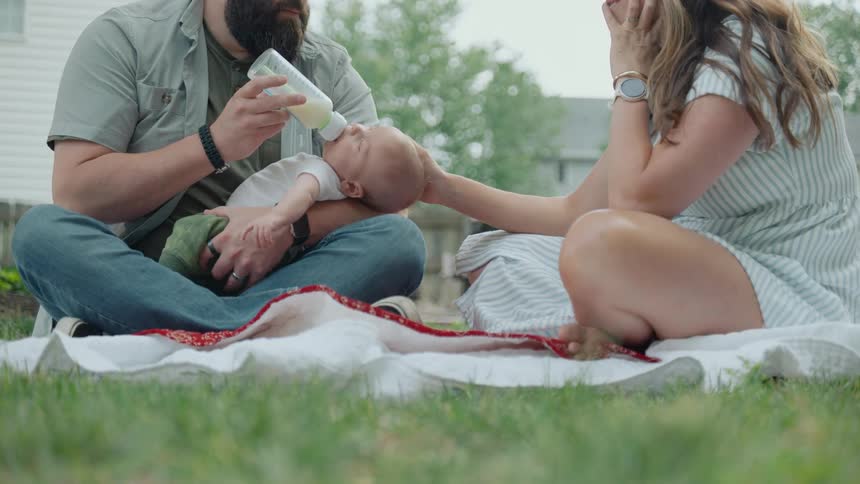 A father holds and feeds his baby with a bottle while the mother lovingly caresses the infant's head on a blanket outdoors