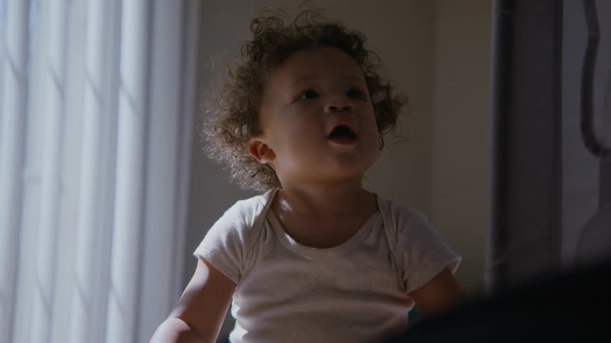 An adorable curly-haired toddler smiles and laughs with wonder while sitting in a softly sunlit room