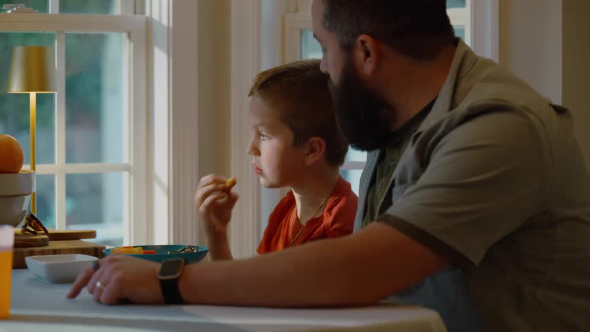 Slow motion side view of a father and son eating snacks together at a dining table by the window
