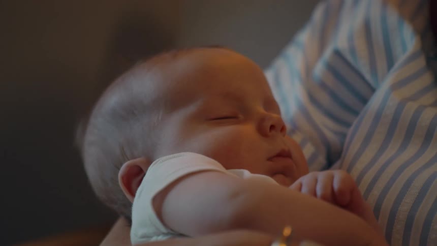A tender close-up shows a newborn baby sleeping peacefully while being held