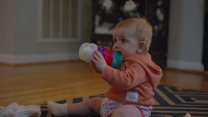 A candid moment of a cute baby girl in an orange hoodie teething on a colorful bottle while sitting on the living room floor