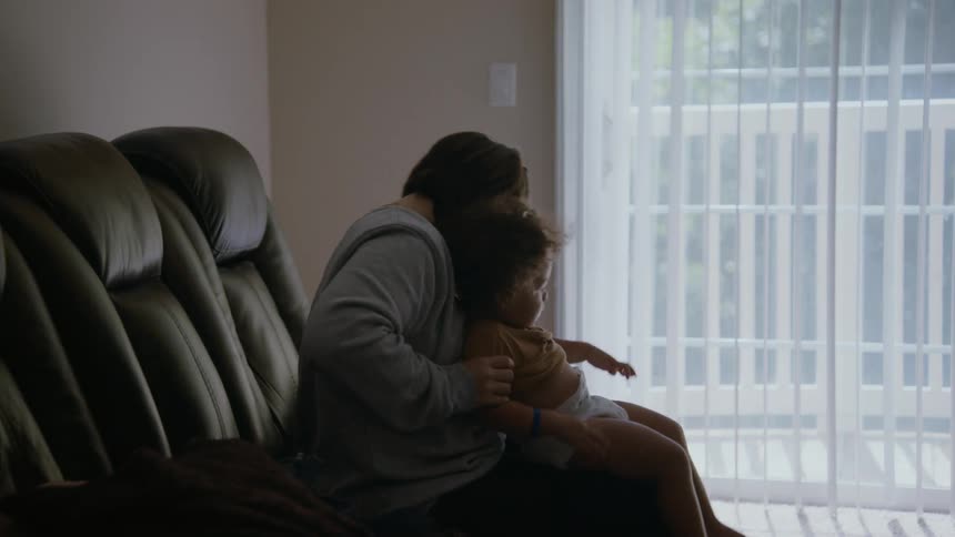 A silhouetted mother gently holds and cuddles her toddler on the couch in a dimly lit room in slow motion
