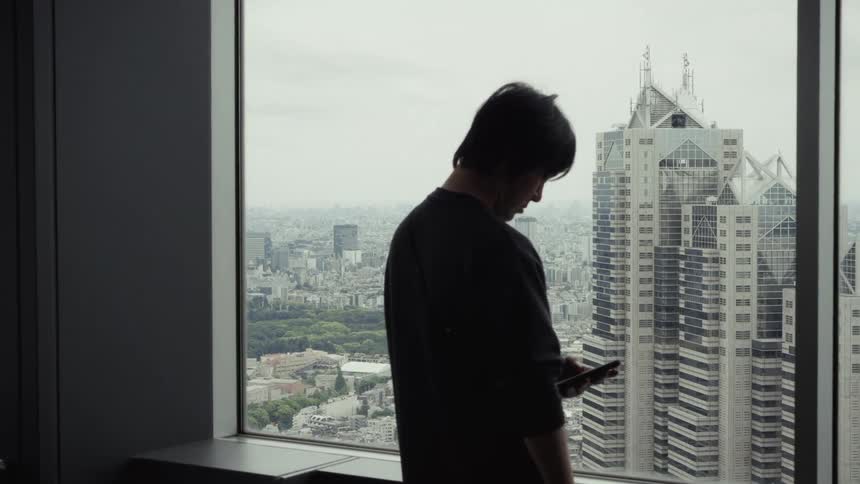 A silhouetted man stands by a high-rise window, looking out over the Tokyo cityscape before checking his phone