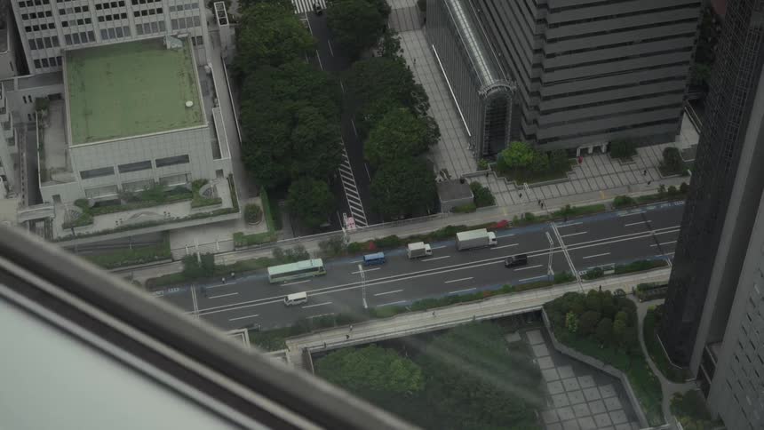 High-angle stationary view from a skyscraper window of traffic moving along a busy, tree-lined street in a modern city