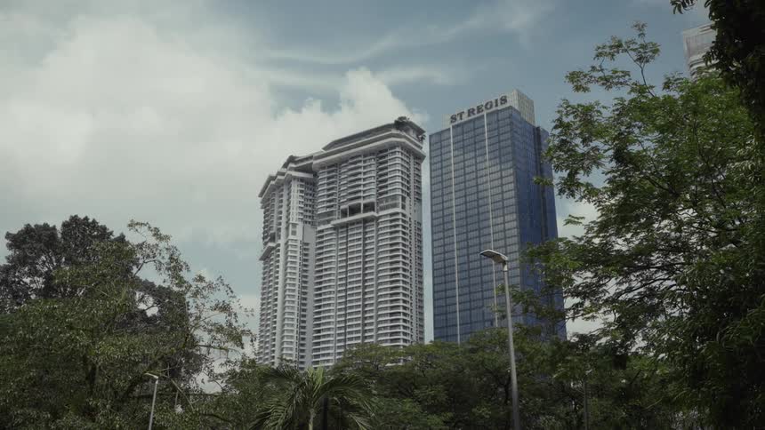 A low-angle view of the modern St. Regis hotel and a neighboring skyscraper seen through lush green trees in Kuala Lumpur