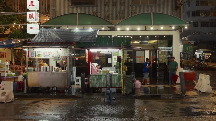 A street food vendor works at their brightly lit stall on a wet city street at night in Malaysia