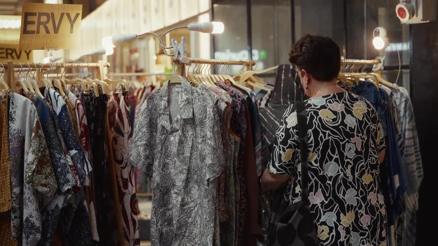 A woman seen from behind browses through a rack of patterned dresses and shirts at a clothing stall in a market