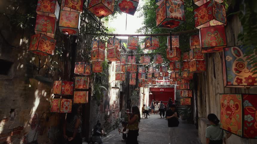 Tourists admire and photograph traditional Asian paper lanterns hanging in a vibrant, narrow city alleyway
