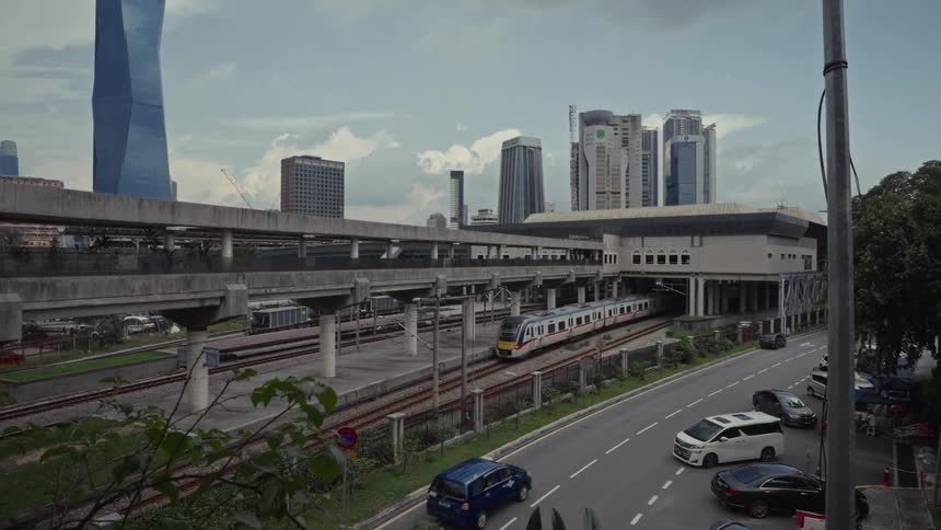 A modern passenger train pulls out of a station in Kuala Lumpur, Malaysia, with the city skyline in the background