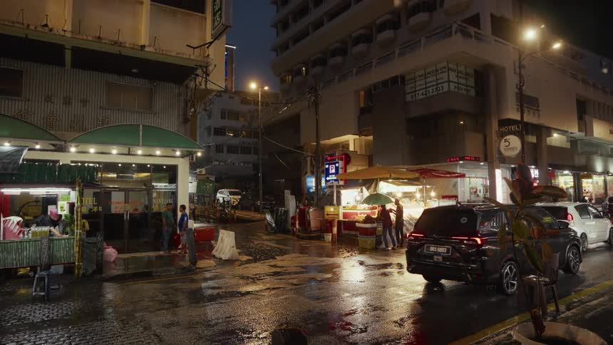 Street food vendors operate on a wet city corner at night as cars slowly pass by in the rain
