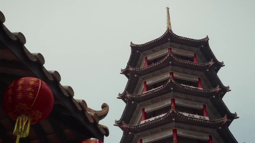 A static, low-angle shot of a traditional Chinese pagoda standing tall against a grey, overcast sky, with a red lantern in the foreground