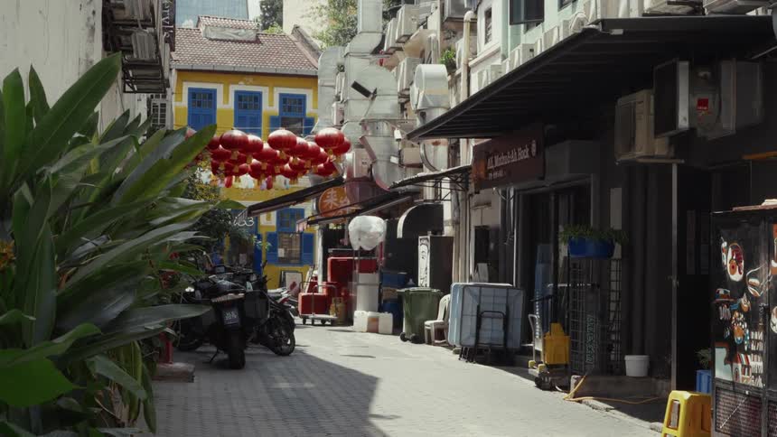 Red Chinese lanterns hang above a quiet, sun-drenched back alley in Singapore, lined with colorful buildings and parked motorcycles