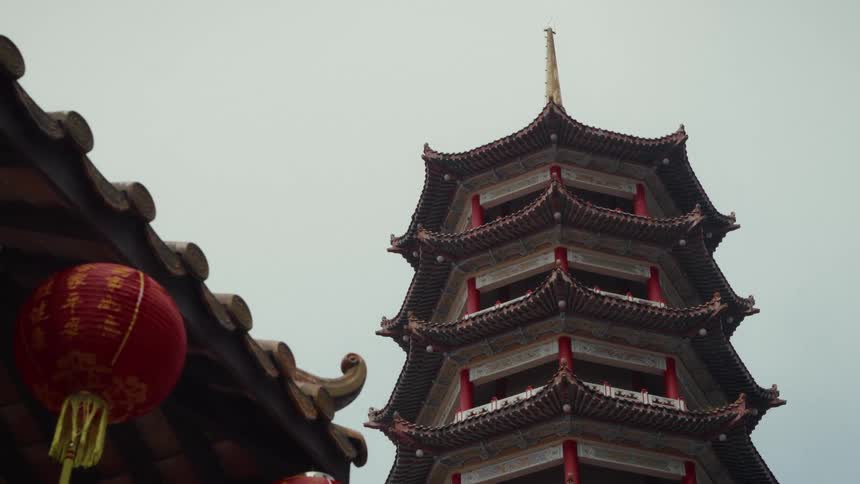 Low angle view of a tall, traditional Chinese pagoda with red paper lanterns hanging in the foreground under an overcast sky