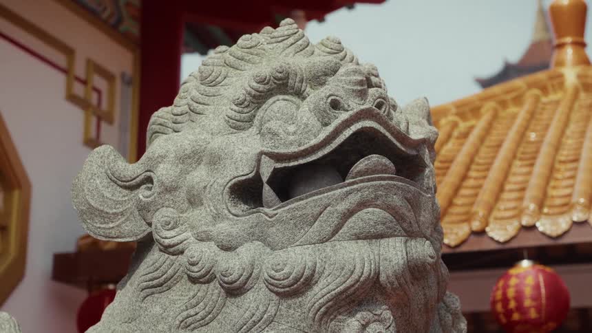 A detailed close-up shot of a traditional Chinese guardian lion statue made of stone, with a temple in the background