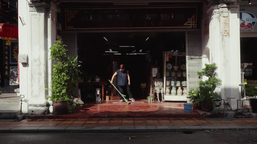 A woman diligently mops the tiled floor at the entrance of her antique shop in Malacca as traffic drives by