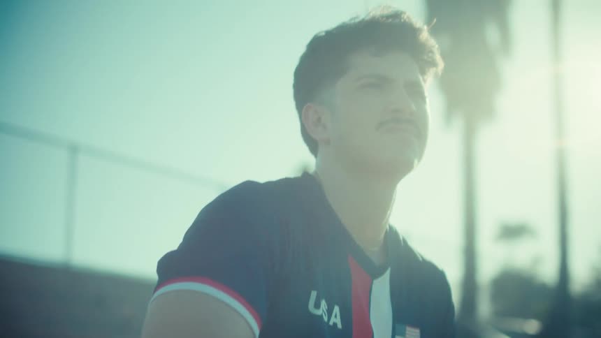 A determined young soccer player in a USA jersey sits on the bleachers with a ball, looking up thoughtfully into the bright sun