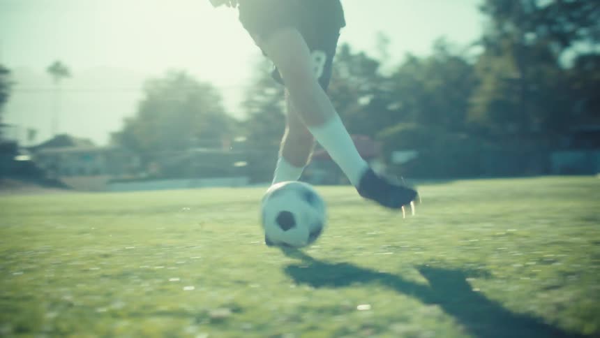 Low angle slow motion shot of a soccer player's feet as he dribbles a ball across a sunny field