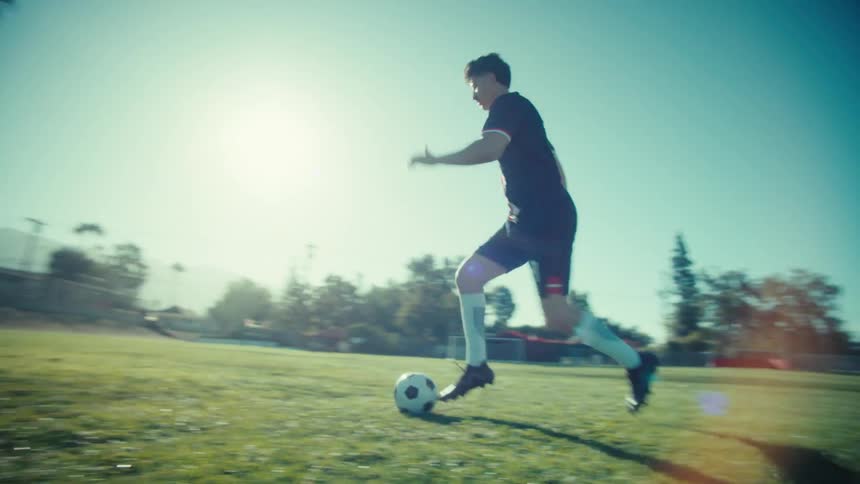 Dynamic slow-motion tracking shot of a young man dribbling a soccer ball across a sunlit field
