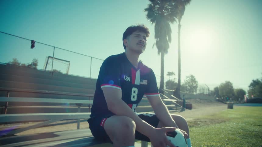 A determined young male soccer player in a USA jersey sits on a bench, contemplating the game in the bright sunshine