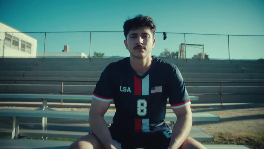 A determined young soccer player in a USA jersey sits on the bleachers holding a soccer ball and looks seriously into the camera