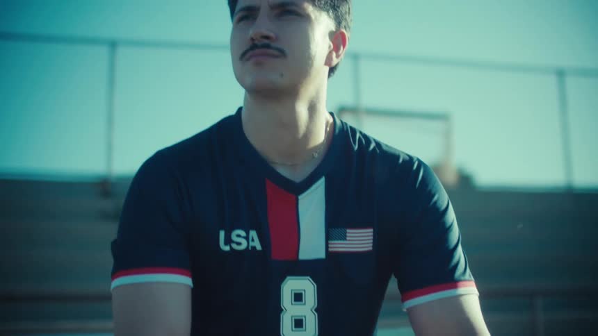 A determined young USA soccer player with a mustache sits on the bleachers holding a soccer ball, gazing into the distance before a game