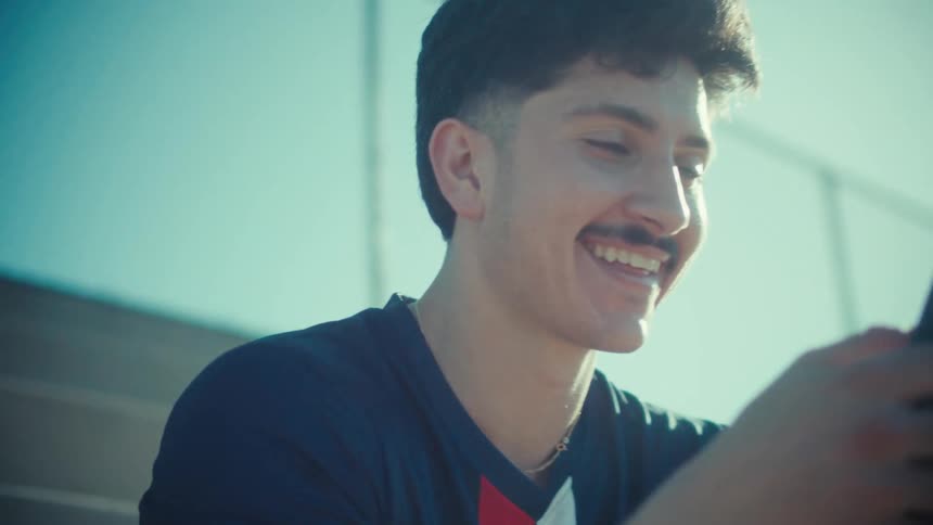 A smiling young man in a sports jersey texts on his phone while sitting on stadium bleachers on a sunny day
