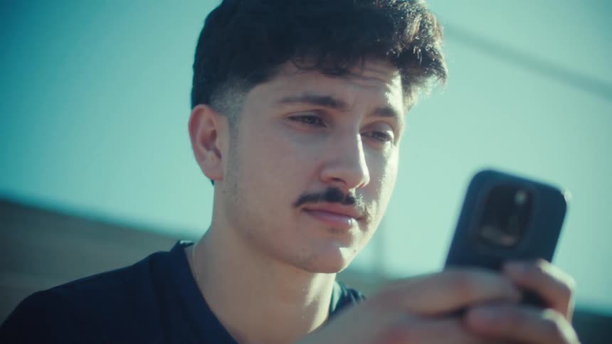 A young male athlete with a mustache sits on bleachers under the bright sun, intently focused on his smartphone
