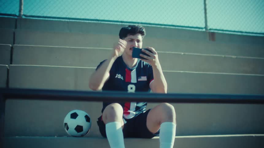A soccer player sits on the bleachers and enthusiastically cheers while watching a game on his smartphone