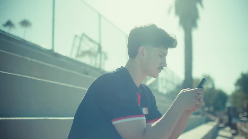 A young male soccer player in uniform sits on concrete stadium bleachers and scrolls on his phone on a bright sunny day
