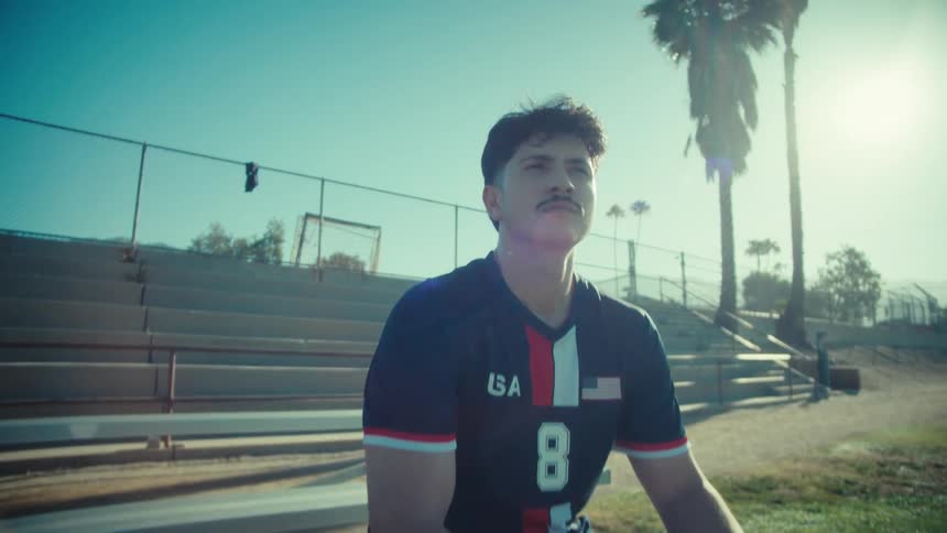 A pensive young soccer player in a USA jersey sits on the bleachers holding a soccer ball on a sunny day