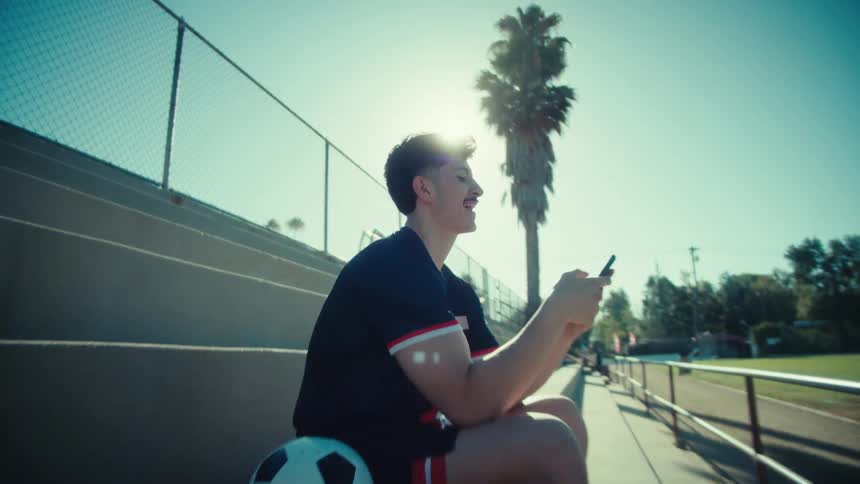 A young soccer player smiles as he texts on his smartphone while sitting on stadium bleachers on a bright, sunny day