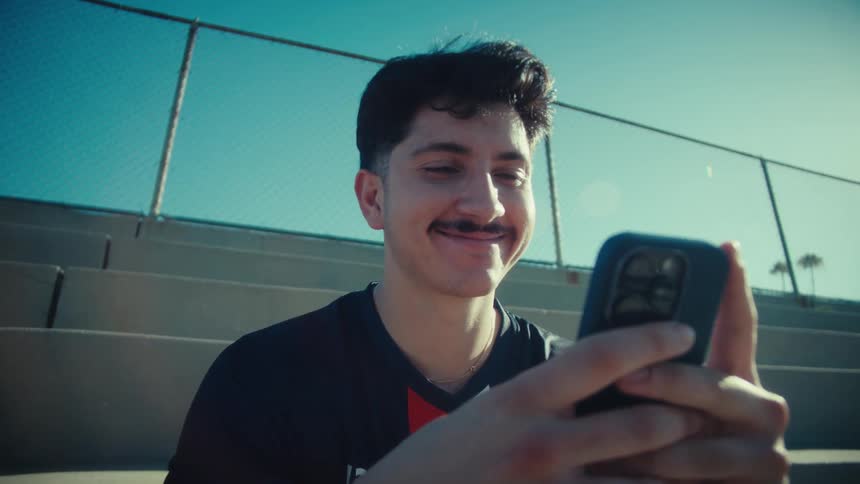 A handsome young athlete with a mustache smiles and laughs while scrolling on his smartphone, sitting on stadium bleachers on a sunny day