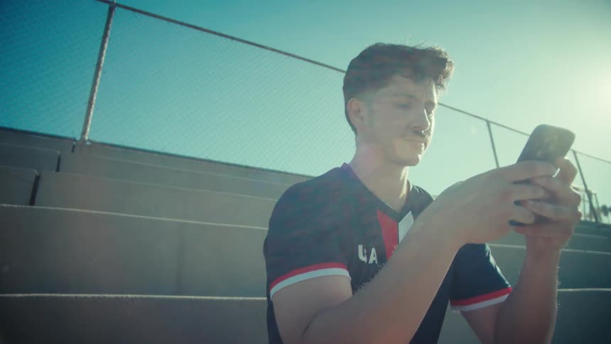 A young soccer player in uniform scrolls on his phone while sitting on sunny stadium bleachers, with the camera slowly pushing in for a close-up