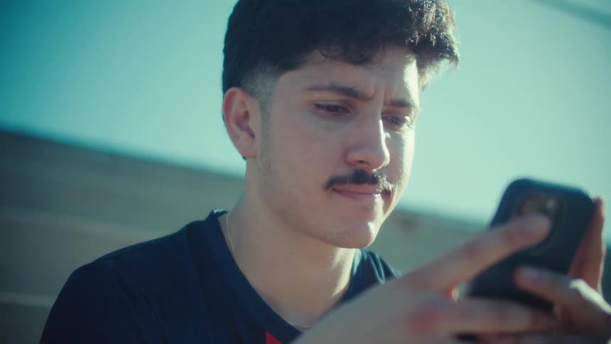 A young man with a mustache sits on the bleachers in the sun, intensely focused on his smartphone