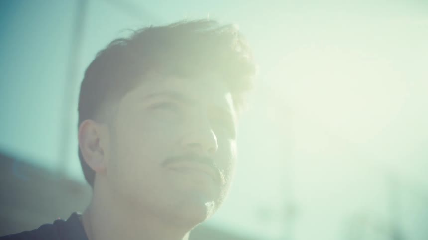 Close up of a young man with a mustache looking up thoughtfully while using his phone in the hazy glow of the bright sun