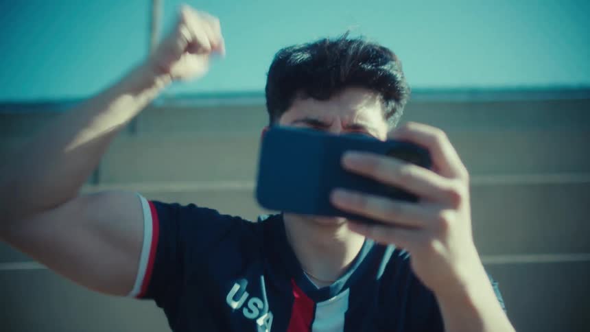 An excited young man with a mustache celebrates winning a mobile game with a triumphant fist pump outdoors