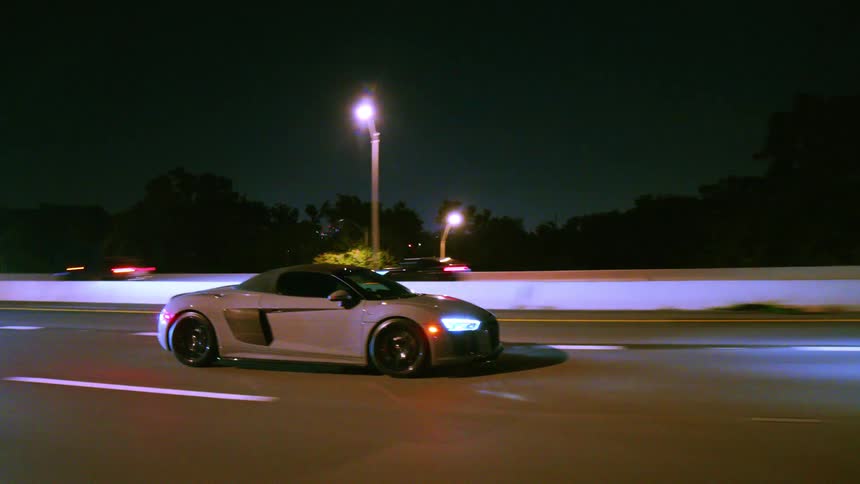 A tracking shot follows a sleek, grey convertible sports car as it cruises down a brightly lit highway at night