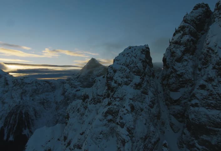 A dramatic aerial flight over jagged, snow-covered mountain peaks at twilight