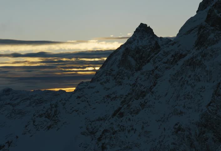 A slow aerial shot reveals a stunning, snow-covered mountain range silhouetted against a golden sunrise sky