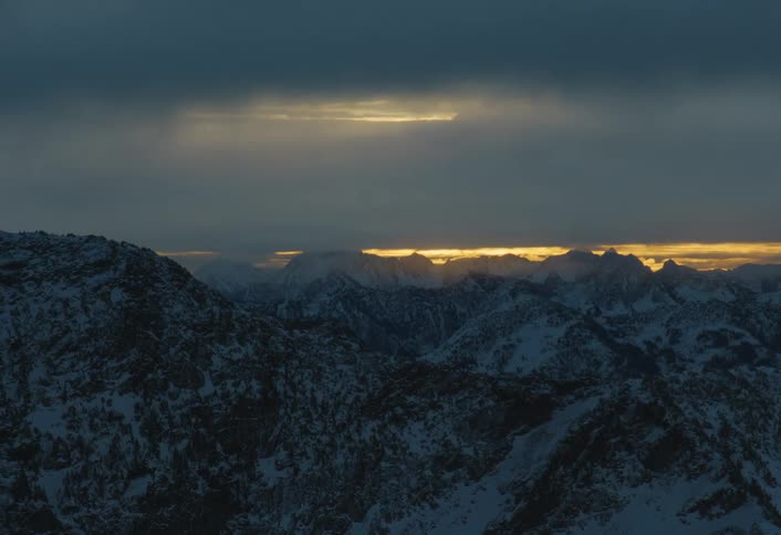 A sliver of golden sunset light breaks through dramatic clouds over a vast, snowy mountain range