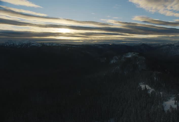 An aerial shot slowly glides over a shadowy, snow-covered forest towards a distant mountain range at sunrise