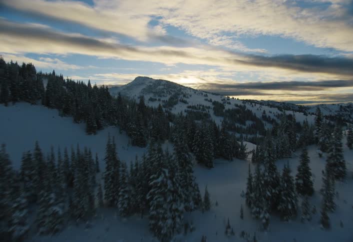A breathtaking aerial shot flies over a serene, snow-covered mountain landscape with evergreen forests at dusk