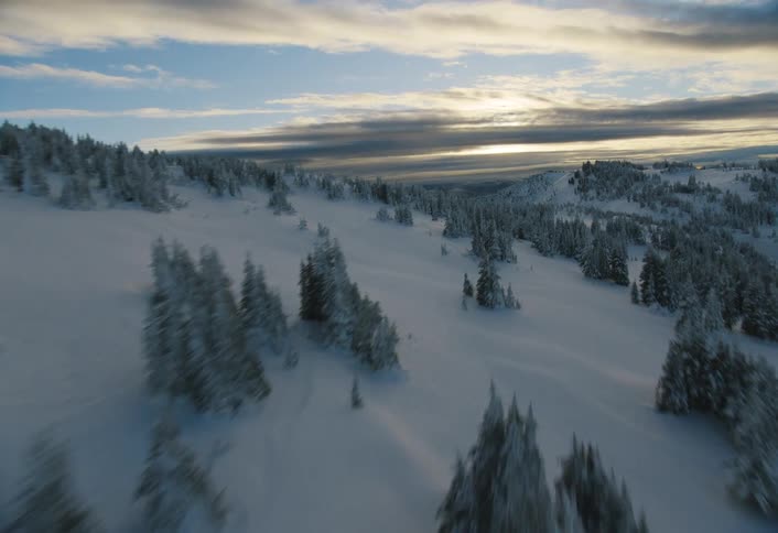 A majestic aerial flight glides over a vast, snow-covered mountain range and pine trees during a beautiful sunset