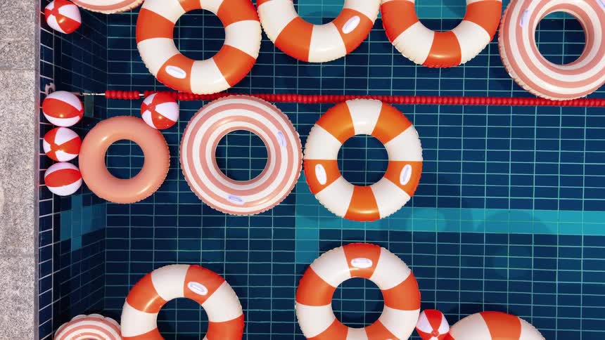 An overhead view of red and white striped inflatable rings and beach balls floating gently in a blue tiled swimming pool