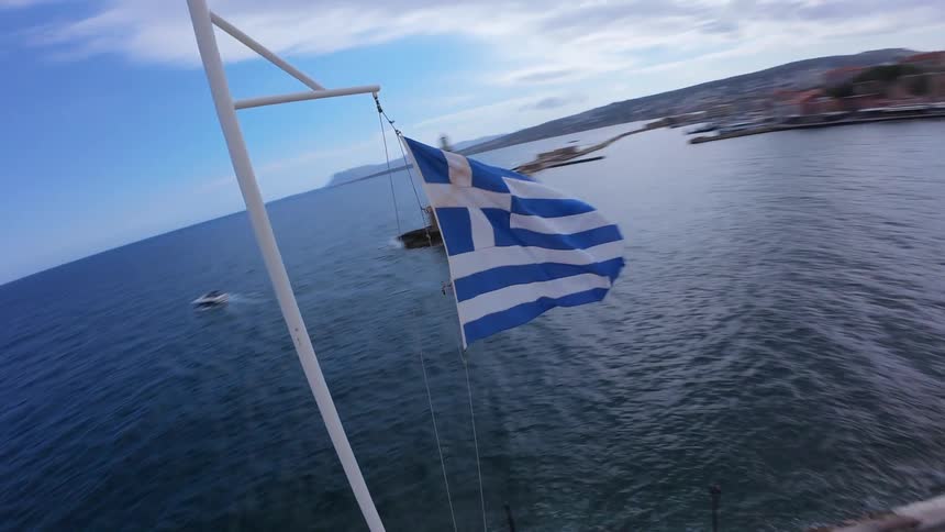 A dynamic, orbiting shot captures the Greek flag waving proudly over the historic harbor town of Chania, Crete, revealing the vast blue sea and distant lighthouse
