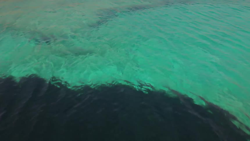 An aerial view flies over crystal clear turquoise water to reveal a luxury resort with private sun decks on a rocky coastline