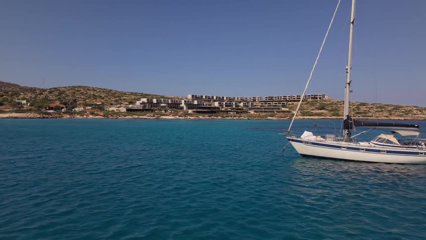 Aerial view of a sailboat anchored in a tranquil turquoise bay with a modern coastal resort in the background